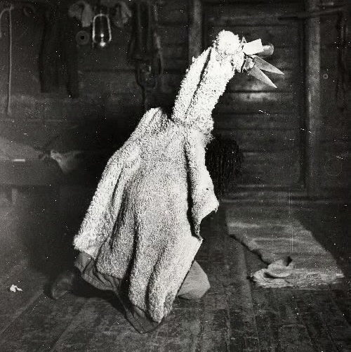 A black and white photo of a person in an old Finnish home, dressed up in white fur and wearing a pointy mask. Illustrating the Kekri harvest festival experience.