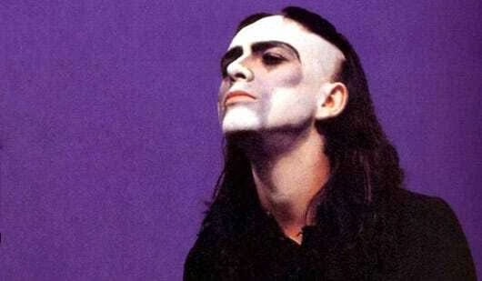 A photo of Peter Gabriel in a black shirt, with white face makeup and dark purple colour over his cheeks, against a purple background.